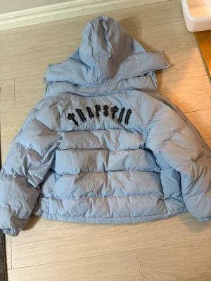 Trapstar ljusblå pufferjacka med huva - Säljer en ljusblå pufferjacka från Trapstar med stor huva och fet logga broderad på ryggen. Jackan har dragkedja framtill, två fickor med knapp och en rund patch på bröstet. Perfekt för kalla dagar och riktigt snygg streetstil.
