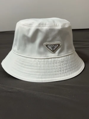 Vit bucket hat från Prada - Säljer en clean vit bucket hat från Prada med ikonisk metallogo framtill. Hatten är tillverkad i polyester med bomullsfoder för extra komfort. Perfekt accessoar för dig som vill ha en stilren och lyxig look. helt ny kommer med lappar och en plastpåse till den tar byten bror på vad och fraktar genom plick 
