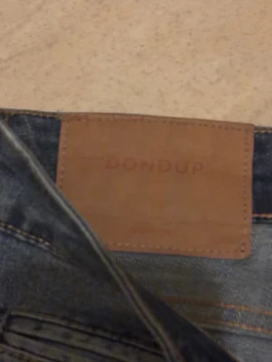 Blå slitna jeans från Dondup George  - Säljer ett par blå jeans från Dondup med snygga slitningar och detaljerade rivna partier. Jeansen har fem fickor, knappgylf och klassisk passform med smalare ben. Materialet är robust denim och färgen är mörkblå med tvättade effekter.