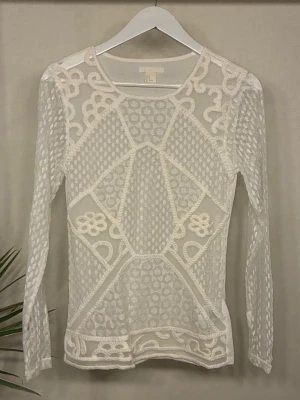Vit spetsblus med mönstrade detaljer - Oanvänd vit långärmad blus med transparent spets och broderade mönster framtill. Toppen har rund halsringning och är figurnära i passformen. Ärmarna är också i spets med diskreta prickar. Perfekt för dig som gillar unika detaljer