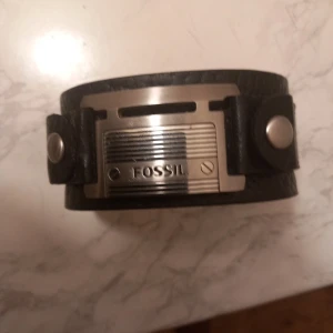 Svart läderarmband från Fossil - Snyggt svart armband från Fossil med bred läderrem och metallplatta framtill. Armbandet har coola detaljer med nitar och ett spänne i silverfärgad metall. Perfekt accessoar för dig som gillar en lite tuffare stil.