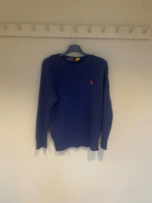 Marinblå stickad tröja Polo Ralph Lauren - Snygg marinblå stickad tröja från Polo Ralph Lauren i storlek S. Tröjan har ett klassiskt kabelstickat mönster och en röd broderad logga på bröstet. Rund hals och långa ärmar, perfekt för kyliga dagar. Passar dig som gillar stilren och tidlös design.