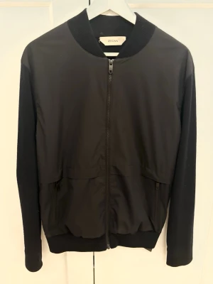 Svart bomberjacka från Zegna - Svart bomberjacka från Zegna med stilren design och ribbad krage. Jackan har dragkedja framtill, två fickor med dragkedja och långärmade ärmar. Materialet är en mix av polyester och stickad textil, vilket ger en snygg kontrast mellan fram- och baksida.