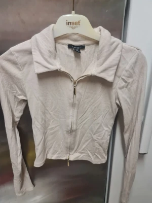 Beige ribbad ziptröja från Amisu - Säljer en beige ribbad tröja från Amisu i storlek XS. Tröjan har lång ärm, krage och snygg dragkedja framtill. Materialet är mjukt och stretchigt, perfekt för lager på lager. Passformen är normal och färgen är ljusbeige.