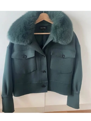 Grön jacka med päls från Meotine - Snygg mörkgrön jacka från Meotine med stor fluffig päls på kragen. Jackan har två stora fickor framtill, knappar och en croppad passform. Perfekt för dig som vill sticka ut med en cool och trendig look. (Knappen längst ner är borta)