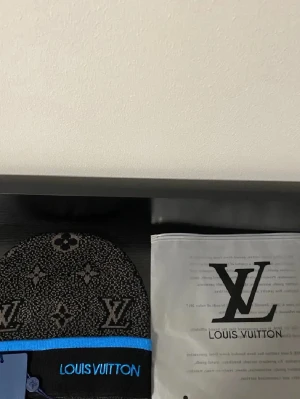 svart mönstrad mössa - Svart mössa med grått monogrammönster och en blå kant längst ner. Märkets logga är broderad i blått framtill. Mössan är stickad och har en klassisk form som passar perfekt för kyliga dagar.