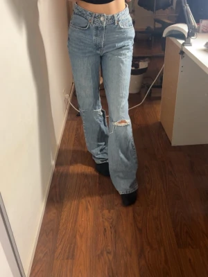 Ljusblå bootcut jeans med slitning - Säljer ett par ljusblå jeans med bootcut-snitt och hög midja. Jeansen har en snygg slitning vid knät. 