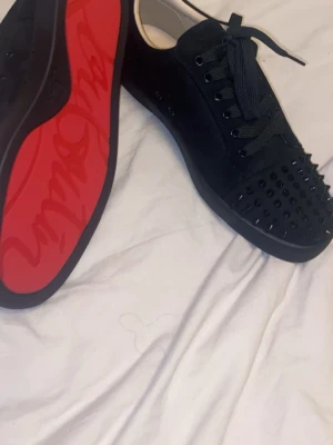 Christian Louboutin svarta sneakers med nitar - Svarta sneakers från Christian Louboutin med ikonisk röd sula och svarta nitar på tån. Skorna har snörning och är tillverkade i mocka med insida i skinn. Snygg och edgy design som sticker ut med detaljer och exklusiv känsla.