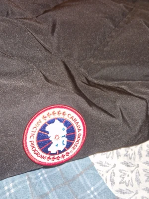 Svart pufferjacka från Canada Goose - Svart pufferjacka från Canada Goose med huva och klassisk logga på ärmen. Jackan har dragkedja och knappar framtill, två stora fickor och quiltad design. Materialet är polyester och dun, perfekt för kalla dagar. Snygg och funktionell för dig som vill ha värme och stil. Jackan släpper ganska mycket fjädrar på insidan och är lite sönder på högersidan.