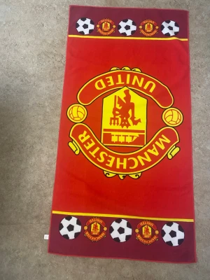 Manchester United badhandduk röd - Stor badhandduk med Manchester Uniteds ikoniska klubbmärke i gult och rött. Handduken har fotbollsmotiv och klubbens logga längs kanterna. Perfekt för stranden eller gymmet för dig som älskar fotboll och vill visa ditt lag.