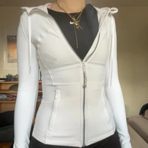 Vit hoodie från Lululemon med dragkedja - Supersnygg vit hoodie från Lululemon med figurnära passform, långa ärmar med tumhål och dragkedja framtill. Tröjan har huva med snörning och diskret logga på dragkedjan. Perfekt för träning eller chill dagar. Tröja har en fläck som man kan se på bilden men för mig har det inte varit något som har stört mig eftersom den inte syns så farligt som man kan tro. Jag säljer den då den är lite för lite för mig.