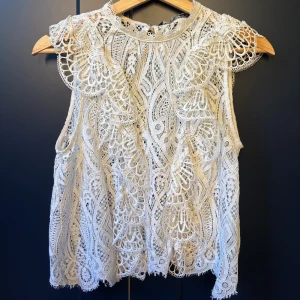Vit spetsblus från Zara - Superfin vit blus från Zara i spets med vackra broderade detaljer och ärmlös design. Blusen har hög halsringning och knäppning bak i nacken. 
