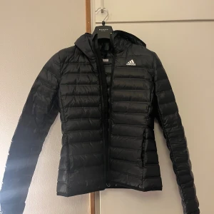 Svart dunjacka från Adidas - Svart dunjacka från Adidas med huva och vit logga på bröstet. Säljer den då den är för liten, endast använd en höst så den är i väldigt fint skick💕