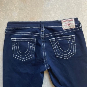 True Religion mörkblå bootcut jeans - Säljer ett par mörkblå jeans från True Religion med tydliga vita kontrastsömmar och ikoniska bakfickor. Modellen är bootcut med låg midja och klassisk femficksdesign. Jeansen är tillverkade i stretchigt denim för en snygg passform. Storlek w29 men lite stora på mig på sista bilden, endast använda en gång men måste sälja då dom är för stora! Nypris 1599. Priset är fast