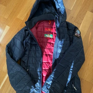 Mörkblå Parajumpers pufferjacka - Snygg mörkblå pufferjacka från Parajumpers. Storlek L men sitter som en S.😆med röd insida och coolt avtagbart patch på ärmen. Jackan har huva, dragkedja och quiltad design. Perfekt för kalla dagar och har en lätt, sportig känsla.