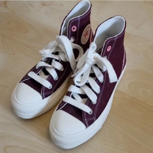 Oanvända vinröda Converse All Star high tops - Snygga vinröda Converse All Star high tops med vita snören och tåhätta. Skorna har rosa detaljer och klassisk logga på sidan. Ovandelen är i canvas och sulan är vit i en platåmodell med rosa undersida. Endast provade en gång! I mycket bra skick förutom några små smutsmärken här och där som knappt syns. Nypris över 600 kr!