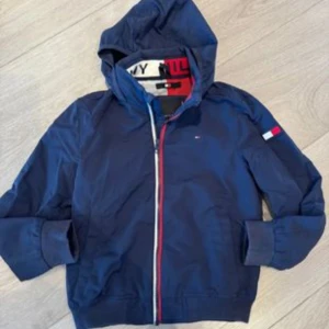 Marinblå vindjacka Tommy Hilfiger - Snygg marinblå vindjacka från Tommy Hilfiger med huva och dragkedja i rött, vitt och blått. Jackan har logotyp på ärmen, ribbade muddar och meshfoder i vitt och rött. Perfekt för dig som gillar klassisk streetstil och vill ha något som sticker ut.