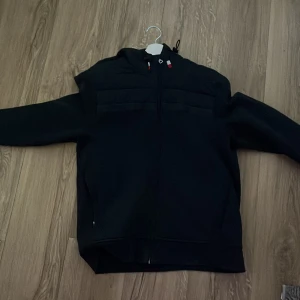 Svart vindjacka från Tommy Hilfiger - Svart vindjacka från Tommy Hilfiger med hel dragkedja, huva med snörning och diskreta röda och vita detaljer. Jackan har quiltade partier upptill och en patch med logga på ärmen. Perfekt för dig som gillar stilren och sportig look.