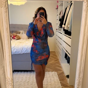 Blå och röd meshklänning med drapering - Säljer en kort, tight klänning i mesh med blått och rött abstrakt mönster. Klänningen har lång ärm, draperad v-ringning och snörning i sidan. Sitter snyggt figurnära och är perfekt för dig som gillar färg och unika detaljer.