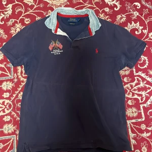 Polo Ralph Lauren piké  - Mörkblå kortärmad piké från Polo Ralph Lauren med broderade amerikanska flaggor och röd logga på bröstet. Skjortan har ljus denimkrage med röd insida och är i slim fit-modell. Perfekt för dig som gillar klassisk amerikansk stil.