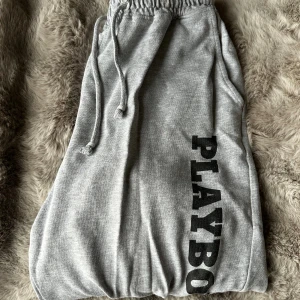 Grå mjukisbyxor från Playboy - Grå mjukisbyxor från Playboy med svart logga längs ena benet. Byxorna har resår och snörning i midjan samt muddar vid bensluten. Fint skick men använda och kan därför finnas någon noppra