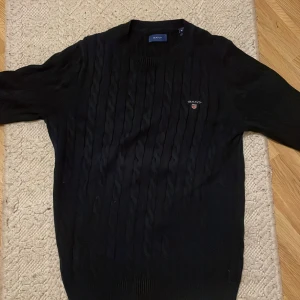 Svart kabelstickad tröja från GANT - Kabelstickad svart tröja från GANT med broderad logga på bröstet. Klassisk rund halsringning och ribbade muddar. Perfekt för dig som gillar stilren och tidlös design. Materialet är mjukt och skönt, passar till många outfits.