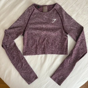 Vinröd croppad träningstopp från Gymshark - Supersnygg vinröd croppad långärmad träningstopp från Gymshark med diskret mönster och logga på bröstet. Toppen har slim passform och är tillverkad i mjuk och stretchig polyester, perfekt för gymmet eller sport.