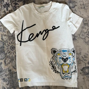 Vit Kenzo t-shirt med tigertryck - Snygg vit t-shirt från Kenzo med stort svart Kenzo-tryck över bröstet och en färgglad tiger på sidan. Klassisk passform och rund hals. Tillverkad i mjuk bomull, perfekt för en cool och avslappnad stil.