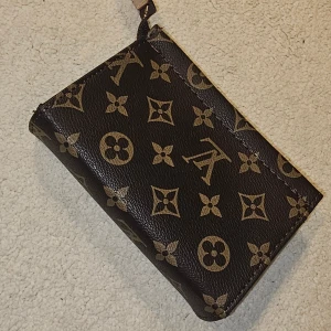 monogram kuvertväska - Snygg kuvertväska från xxxx med klassiskt monogrammönster i brunt och beige. Väskan är tillverkad i skinn och har dragkedja upptill. Perfekt storlek för det viktigaste och en ikonisk accessoar med lyxig känsla.Betala med direkt swish jag kan salja for 650kr plus frakt