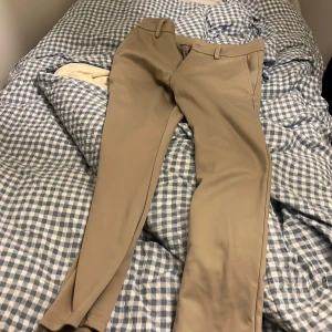 Beige kostymbyxor från Twosociety - Snygga beige kostymbyxor från Teshop med raka ben och klassiska fickor både fram och bak. Byxorna har hällor för bälte och stängs med knapp och dragkedja. Perfekta för en clean och stilren look.