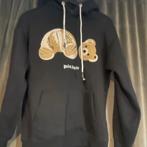 Svart Palm Angels hoodie med björn - Svart hoodie från Palm Angels med ikoniskt björnmotiv framtill. Tröjan har huva med vita snören och stor magficka. Mjuk insida och klassisk passform, perfekt för dig som gillar streetwear och vill sticka ut med en unik detalj. Passar också för dem i storlek M