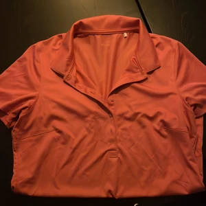 Korallrosa piké från Puma - Sportig korallrosa pikétröja från Puma med klassisk krage och knappar framtill. Tillverkad i ett lätt och svalt syntetmaterial som passar perfekt för aktiva dagar. Kortärmad och stilren design som ger en fräsch look.