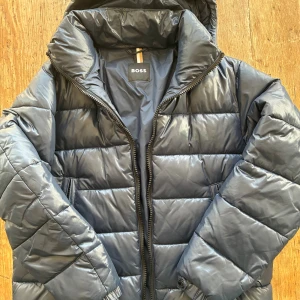 Mörkblå pufferjacka från BOSS - Snygg mörkblå pufferjacka från BOSS med huva och dragkedja. Jackan har quiltad design, två fickor med dragkedja och en stilren look. Den är i princip i nyskick. Nypris runt 4000. Kan lösa kvitto vid efterfrågan