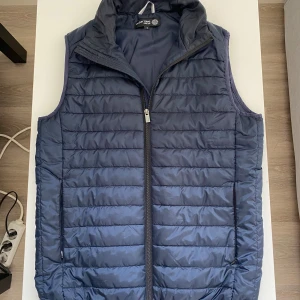 Blå dunväst från On Our Terms - Snygg blå dunväst från On Our Terms med quiltad design och dragkedja framtill. Västen har ståkrage och två sidofickor med dragkedja. Perfekt för lager-på-lager och enkel att matcha med hoodies eller tröjor.