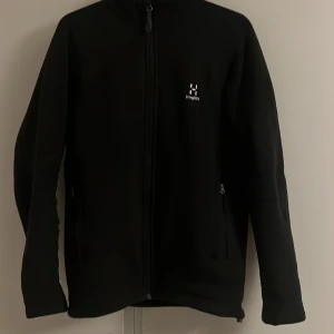 Svart fleecejacka från Haglöfs, S - Svart fleecejacka från Haglöfs i storlek S. Jackan har hel dragkedja framtill, två sidofickor med dragkedja och justerbar nederkant med snörning. Perfekt för kyliga dagar och outdoor-häng. Klassisk och stilren design med Haglöfs-logga på bröstet.