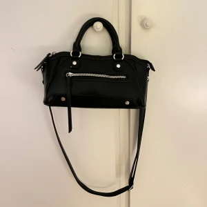 Svart handväska med dragkedja - Snygg svart handväska i skinnimitation med både handtag och lång axelrem. Väskan har silverfärgade detaljer och en dragkedja framtill. 