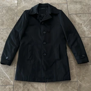 Svart rock från Selected Homme - Stilren svart rock från Selected Homme med klassisk krage och knäppning framtill. Jackan har två sidofickor, knappar vid ärmslut och quiltat foder på insidan för extra värme. Perfekt för dig som vill ha en clean och tidlös look.