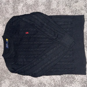 Svart kabelstickad tröja Polo Ralph Lauren - Kabelstickad svart tröja från Polo Ralph Lauren med klassisk röd broderad logga på bröstet. Tröjan har rund halsringning, ribbade muddar och lång ärm. Perfekt för dig som gillar stilren och tidlös design.