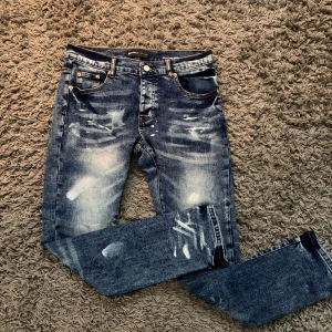 Blå distressed jeans från Purple Brand - Säljer ett par blå jeans från Purple Brand med coolt distressed och blekt mönster. Jeansen har klassisk femficksdesign, orangea sömmar och snygga slitningar framtill och baktill. Perfekt för dig som gillar en edgy streetstil.