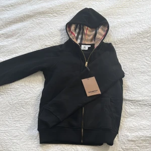 Svart Burberry hoodie med dragkedja - Svart hoodie från Burberry med klassiskt rutigt foder i huvan och guldfärgad dragkedja. Storlek M men passar mer som S