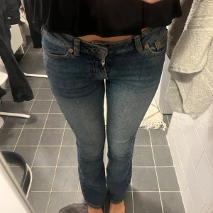  blå bootcut jeans - Säljer ett par klassiska jeans i blå tvätt med bootcut passform. Jeansen har låg midja, fem fickor och snygga kontrastsömmar. Materialet är slitstarkt denim i bomull och de har dragkedja och knapp framtill. Perfekta för dig som gillar retrovibbar och tidlös stil.