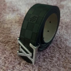 Svart Louis Vuitton bälte med rutigt mönster - Snyggt svart bälte från Louis Vuitton med klassiskt rutigt mönster och en stor silvrig LV-spänne framtill. Bältet är gjort i skinn och har en stilren design som passar till många olika outfits. Perfekt accessoar för att lyfta din look.