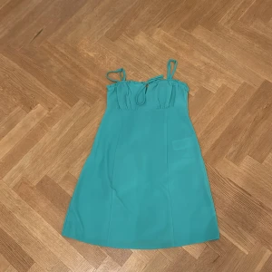 Turkos kort klänning med knyt - Snygg turkos kortklänning med smala axelband och knyt fram upptill. Klänningen har en enkel, rak passform och är perfekt för varma dagar. Materialet känns lätt och svalt, vilket gör den skön att bära under sommaren.