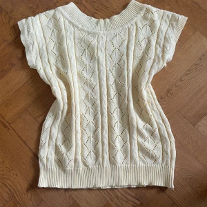 Stickad ärmlös topp i ljusbeige - Supersöt stickad topp i ljusbeige med ärmlös design och bred ribbad kant vid hals och nederdel. Toppen har ett snyggt hålmönster med kabelstickade detaljer och diamantformade partier. Perfekt för lager-på-lager och ger en chill men ändå stilren vibe.
