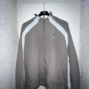 Cortiez jacka - Säljer en grå vindjacka med vita paneler längs ärmarna och dragkedja framtill. Jackan har huva med snörning och en liten logga på bröstet. Perfekt för dig som gillar sportig och clean stil.