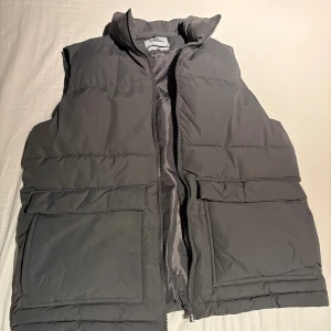 Grå dunväst - Säljer en grå dunväst från Garment med hög krage och två stora fickor framtill. Västen har quiltad design och stängs med dragkedja. Perfekt att slänga över en hoodie för extra värme under kyliga dagar.