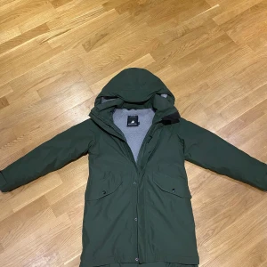 Grön jacka från Didriksons med huva - Säljer en mörkgrön parkas från Didriksons med mysigt grått fleecefoder och stor huva. Jackan är 100% vattentät, har stora fickor med lock och stängs med både dragkedja och knappar. Perfekt för kalla och regniga dagar.