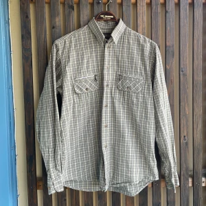 Rutig grön skjorta - Klassisk rutig skjorta från Tuxer Adventure i beige och grå toner. Skjortan har två bröstfickor med kedja och knappar, lång ärm och rak passform. Perfekt för dig som gillar en avslappnad och tidlös stil.