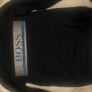 Svart långärmad tröja Hugo Boss - Svart långärmad tröja från Hugo Boss med stor blå och vit logga framtill. 