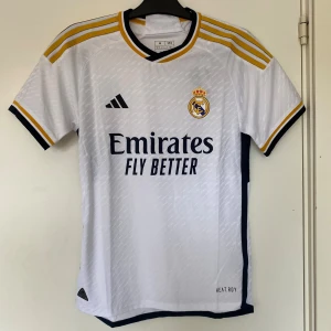 Real Madrid hemmatröja Adidas M - Snygg Real Madrid hemmatröja från Adidas i slim fit, tillverkad i återvunnen polyester. Tröjan är vit med gula och mörkblå detaljer, klassiska Adidas-ränder på axlarna och klubbmärke på bröstet. HEAT.RDY-teknologi för extra komfort.